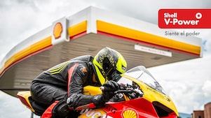 Motociclista con casco y traje de carreras sobre una moto deportiva, frente a una estación de servicio Shell. En la parte derecha de la imagen aparece el logotipo de Shell V-Power y el texto “Siente el poder”.