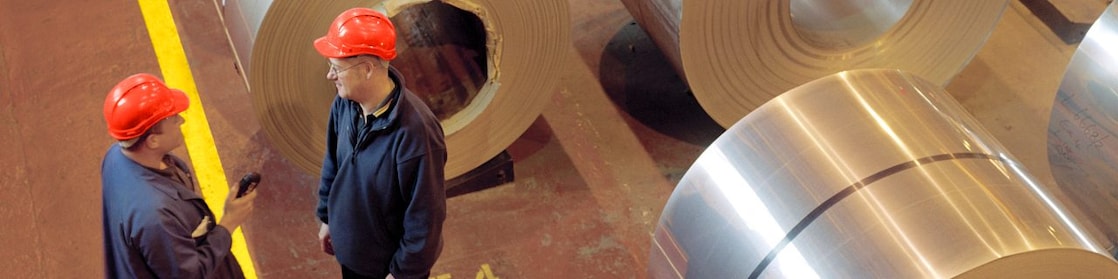Two factory workers talking next to large rolls of steel