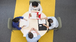 Cuatro empresarios sentados alrededor de una mesa redonda leyendo documentos, alfombra gris y amarilla