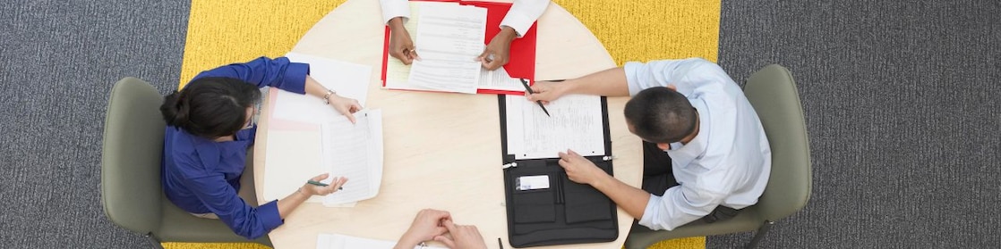 Cuatro empresarios sentados alrededor de una mesa redonda leyendo documentos, alfombra gris y amarilla